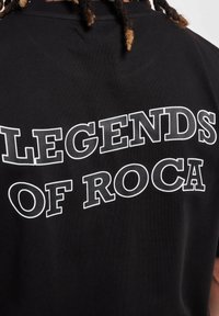 Juodas marškinėliai su ryškiu baltu užrašu "ROCA LEGENDOS" ant nugaros. Medvilnės audinys su lygiaTextūra.