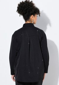 Camicia nera con puntini bianchi, maniche lunghe, colletto e piega posteriore. Realizzata in un tessuto morbido con una leggera lucentezza.