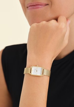 Montre avec un bracelet en maille dorée et un cadran rectangulaire blanc, comportant de simples index horaires et une petite couronne, portée sur un poignet à teint de peau naturel.
