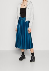 Felpa grigia cropped con zip frontale, top nero senza maniche, gonna midi plissettata turchese, calzini neri e sneakers nere completano l'outfit.