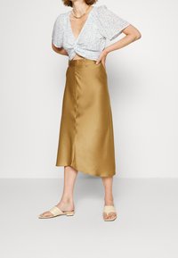 Blusa de estampado floral azul claro con mangas abullonadas, anudada en la cintura; falda midi de satén dorado; sandalias abiertas beige con un sutil relieve.