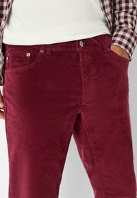 Bordeaux ribfluwelen broek met verticale ribbels, voorzien van zijzakken, metalen knoopsluiting, en gecombineerd met een geruite blouse.