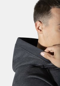Hiilenharmaa huppari, jossa on ribatty kaula-aukko; käsi säätämässä kaulusyksityiskohtia. Pehmeä kangasrunko, minimalismi. Lähikuva kaula-alueesta.