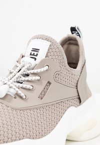 Baskets tricotées beige clair avec une semelle blanche épaisse, des lacets élastiques à motif, et une languette d'extraction portant l'inscription "NEW" au talon.