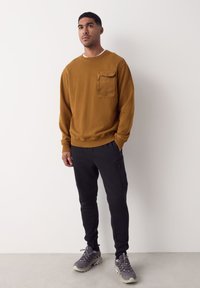 Bruine katoenen trui met een buidelzak aan de voorkant, gecombineerd met zwarte taps toelopende joggers met zijritsen, en grijze chunky sneakers.