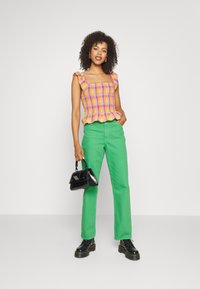 Top de xadrez colorido com alças com pregas, combinado com calças verdes de perna reta e botas de tornozelo pretas segurando uma pequena mala preta.