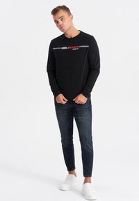Schwarzes Langarmshirt mit Textgrafik; kombiniert mit dunklen, schmal geschnittenen Jeans und weißen Sneakern. Glattes Material, lässige Passform.