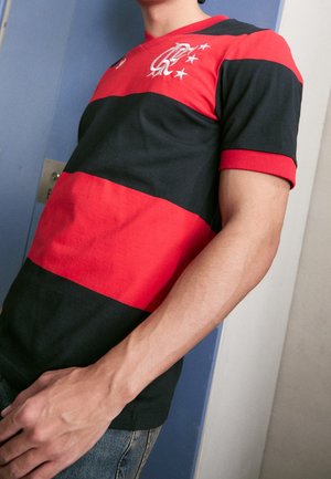 Person trägt ein rot-schwarz gestreiftes Sporttrikot mit einem weißen Logo und vier Sternen auf der Brust und steht drinnen vor blau-weißen Wänden.