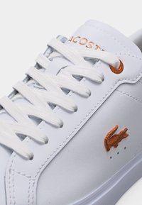 Λευκό δερμάτινο αθλητικό παπούτσι με λευκά κορδόνια, μπρούτζινο λογότυπο Lacoste στη πλευρά και μπρούτζινα ροζέτα κοντά στην κορυφαία τρύπα των κορδονιών.