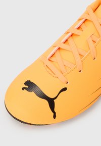 Oranssi jalkapallokenkä sileällä synteettisellä yläosalla, musta Puma-logo ja tonaliset nauhat. Sisältää tekstuuripinnan ja pyöristetyn varren.