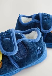Blaue Hausschuhe für Kinder aus plüschigem Stoff, mit Klettverschluss und weicher, weißer Innenausstattung, die auf der Vorderseite ein gelbes Akzentdesign zeigen.