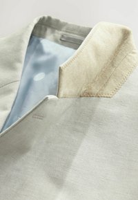 Gros plan sur le col d'une veste gris clair avec un détail en daim beige et une doublure intérieure bleu clair visible avec un motif subtil.