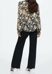 Femme aux cheveux longs portant un chemisier transparent à motifs floraux, un pantalon noir à jambes larges avec des ourlets effilochés, et des sandales blanches à talons, debout de dos face à l'avant.