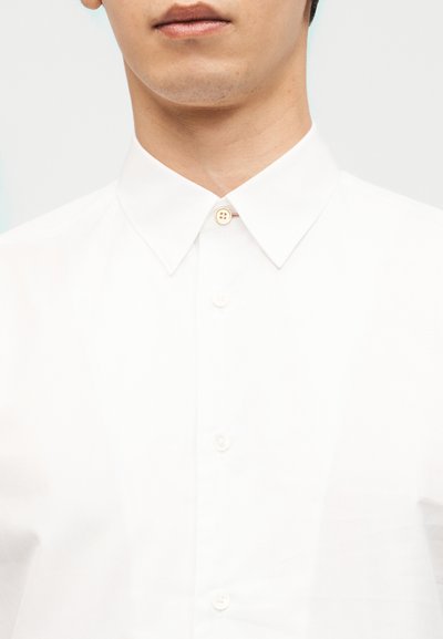 Κοντινό πλάνο ενός ατόμου που φοράει λευκό πουκάμισο με κουμπιά, κλασικό γιακά και κουμπί πάνω κουμπωμένο.