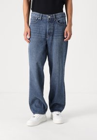 Blugi din denim albastru cu o croială lejeră, cu picioare largi. Au buzunare frontale, închidere cu button și o textură ușor decolorată. Potriviți cu adidași albi.