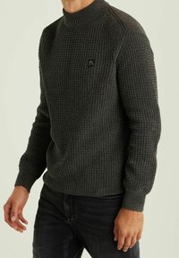 Grauer strukturierter Pullover mit hohem Kragen, gerippten Bündchen und Saum, mit einem schwarzen Logo-Patch auf der Brust. Getragen über dunklen Jeans.