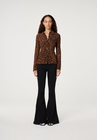 Vrouw staat met een bruin panterprint knoopshirt, zwarte flared broeken en gestreepte schoenen tegen een effen witte achtergrond.
