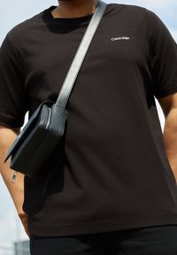 En person som bär en svart Calvin Klein-t-shirt med en mörkgrå axelväska hängande över axeln mot en ljus himmelsbakgrund.