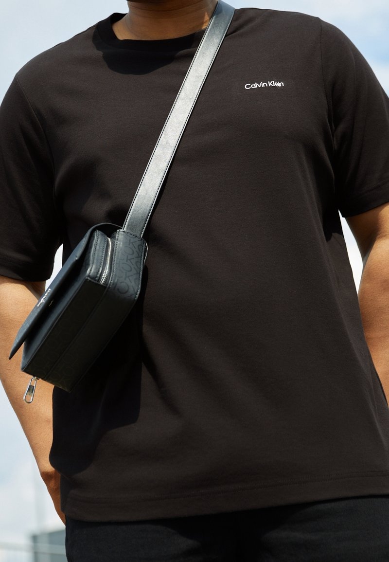 En person som bär en svart Calvin Klein-t-shirt med en mörkgrå axelväska hängande över axeln mot en ljus himmelsbakgrund.