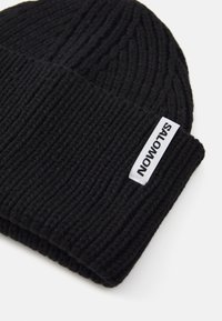 Black knitted beanie hat with a ribbed texture and a folded cuff. Features a white label with black text attached to the side.