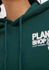 Sudadera de algodón verde con cordones y un logo que presenta el texto "PLAN SHOP" y "PLANT NURSERY" en blanco. Textura suave, diseño casual.