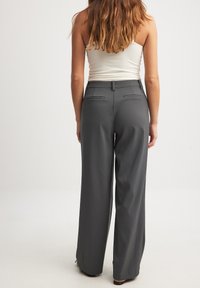 Pelēkas platajās bikses, izgatavotas no gludas auduma, ar jostasvietu, aizmugurējām kabatām un pieguļošu piegriezumu, valkātas ar vienkāršu baltu topu.