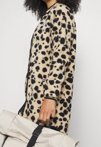 Hřejivý kardigan s leopardím vzorem a černými akcenty; modelka drží světlou šedou bavlněnou tašku s černým popruhem. Kardigan má zipové zapínání.