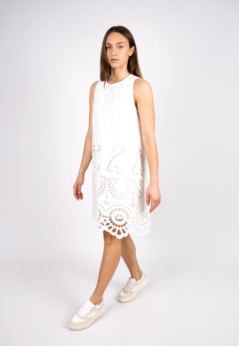 White sleeveless dress with intricate cut-out detailing at the hem, featuring a soft texture and relaxed fit, paired with white sneakers.