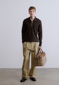 Καφέ πουλόβερ με φερμουάρ, μπεζ παντελόνι με πιέτες, μαύρα παπούτσια και τσάντα ταμπά με καφέ λεπτομέρειες, τοποθετημένα μπροστά σε ουδέτερο φόντο.