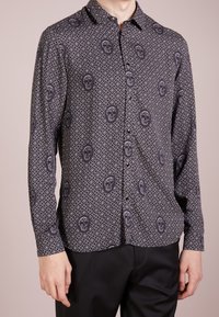 Camicia a maniche lunghe in tessuto scuro con un motivo geometrico a diamante e accenti di teschi in colore più chiaro. Collo a bottoni.