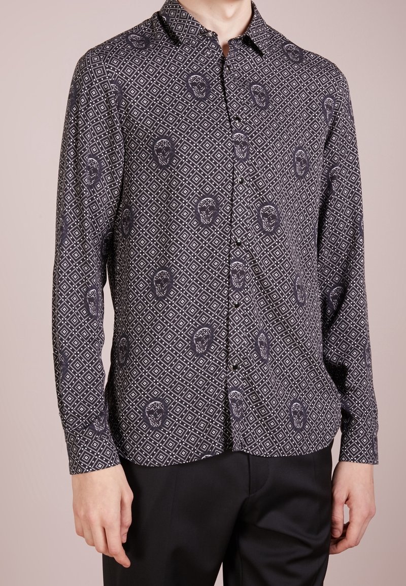 Camicia a maniche lunghe in tessuto scuro con un motivo geometrico a diamante e accenti di teschi in colore più chiaro. Collo a bottoni.
