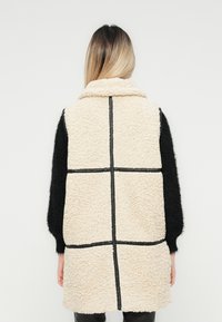 Kvinna med ombre hår som bär en ärmlös beige sherpa-väst med svarta vertikala och horisontella detaljer över svarta långärmade tröjor.