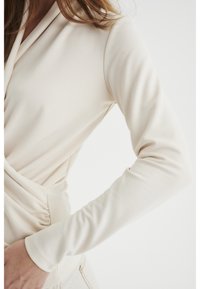 Kvinna som bär en långärmad, krämfärgad wrap-top delvis instoppad i ljusa byxor mot en vit bakgrund.