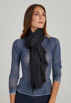 Donna con lunghi capelli castani indossa una maglia azzurra trasparente a maniche lunghe e una sciarpa nera a quadri, guarda leggermente di lato.