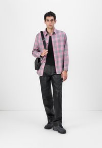 Camisa de botonada a cuadros en rosa y gris, camiseta negra, pantalones de cuero negro brillantes y zapatillas deportivas negras. El modelo lleva un bolso cruzado.