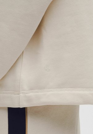 Beżowy rękaw bluzy Lululemon z widocznym logo nad prążkowanym ściągaczem oraz część dopasowanych beżowych spodni z czarnym paskiem po boku.