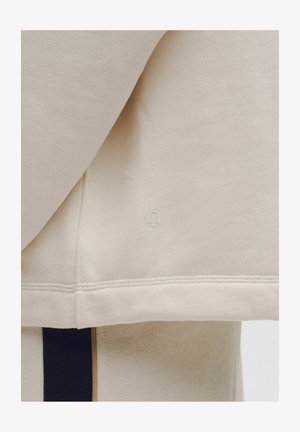 Beżowy rękaw bluzy Lululemon z widocznym logo nad prążkowanym ściągaczem oraz część dopasowanych beżowych spodni z czarnym paskiem po boku.