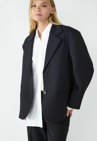 Czarna, oversize marynarka z klapami, dwoma guzikami i kieszeniami z przodu, założona na białą koszulę, o gładkiej teksturze i strukturalnym kroju.