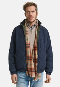 Navy gewatteerde jas met groene kraag, gedragen over een beige sweater en een geruite sjaal met oranje en groene accenten. Gekozen in combinatie met lichtblauwe jeans.