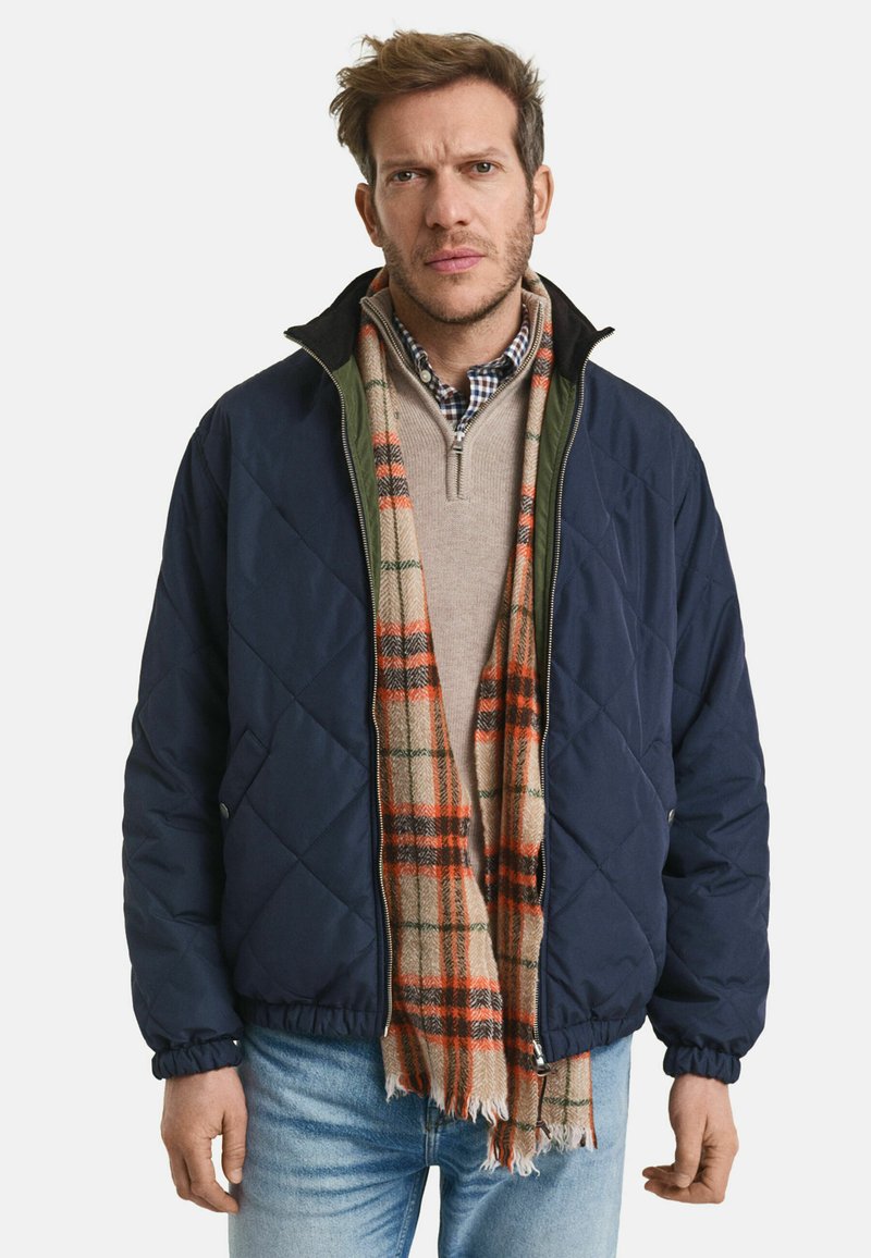 Marineblaue Steppjacke mit grünem Kragen, über einem beigen Pullover und einem karrierten Schal mit orangefarbenen und grünen Akzenten getragen. Kombiniert mit hellblauen Jeans.