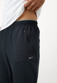 Schwarze Sporthose mit einer Reißverschlusstasche am linken Bein und einem silbernen Nike-Logo. Der Stoff hat eine glatte Textur mit einem leichten Glanz.