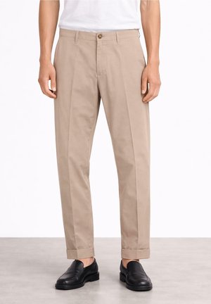 Uomo che indossa pantaloni beige a gamba dritta con orli risvoltati, camicia bianca e scarpe nere in pelle senza lacci, in piedi su un pavimento grigio.