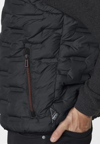Schwarze gepolsterte Weste mit einer strukturierten Strick-Schulter, ausgestattet mit einer Reißverschlusstasche und einem Logo-Patch, sowie einem kontrastierenden orangefarbenen Reißverschlussdetail.