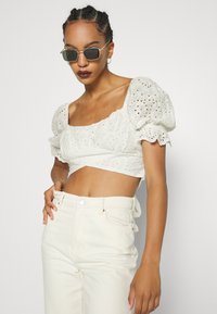 Top branco cropped com mangas bufantes, adornado com bordados em ilhós. Combinado com jeans de cintura alta em tom claro e óculos de sol.