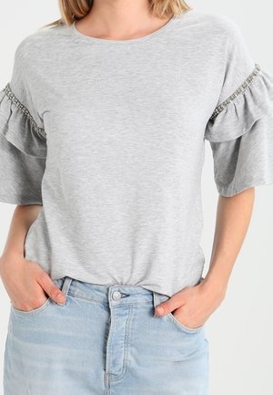 Femme portée un haut gris clair à manches courtes avec des volants et des perles sur les manches, rentrée dans un jean en denim bleu clair avec les mains dans les poches.