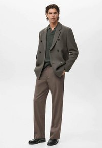 Cappotto grigio doppiopetto sopra una camicia verde scuro, abbinato a pantaloni marroni e scarpe nere, con una vestibilità sartoriale e una texture liscia.