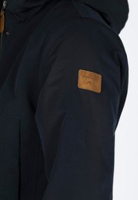 Giacca della marina con cappuccio, realizzata in tessuto liscio, con una toppa in pelle marrone sul braccio superiore. Include una chiusura con zip e una tasca laterale discreta.
