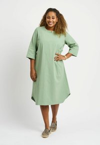 Light green, knee-length dress with three-quarter sleeves, round neckline, and side slits. Paired with leopard print slip-on shoes.