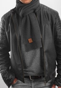 Black leather jacket with quilted shoulder accents, grey knit scarf with a textured pattern, and a tan leather logo patch. Grey sweater underneath.
