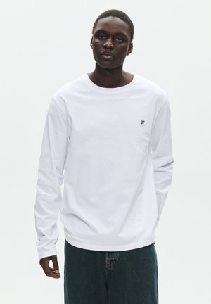 Ung person med kort hår iført en ensfarvet hvid langærmet skjorte med lille logo på brystet og mørkeblå jeans mod en hvid baggrund.